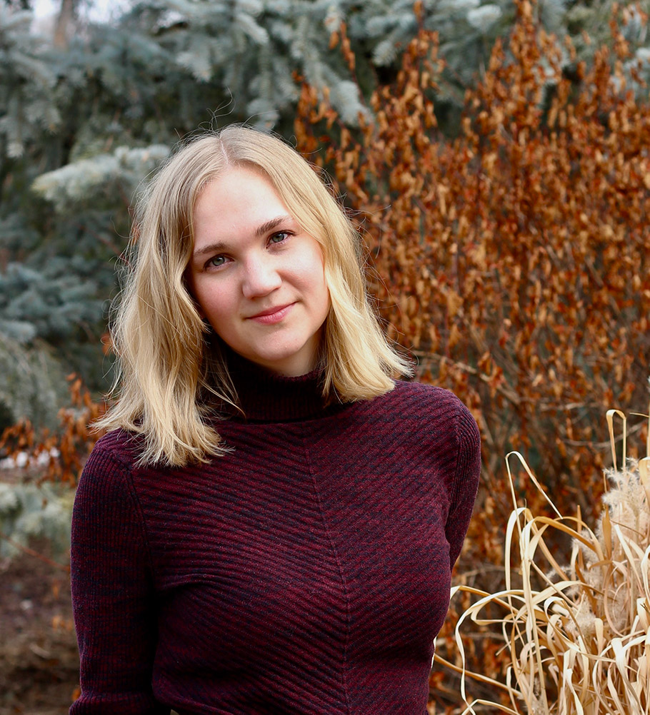 photo: Sarah Ens posing in front of Fall foliage and evergreen trees.